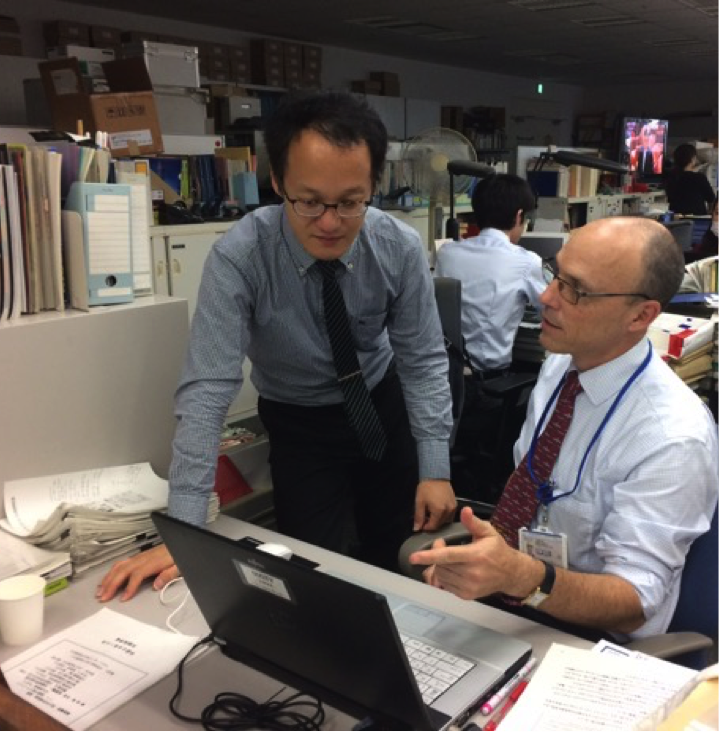 Two male professionals at a desk, one stands over the other sitting in front of a laptop