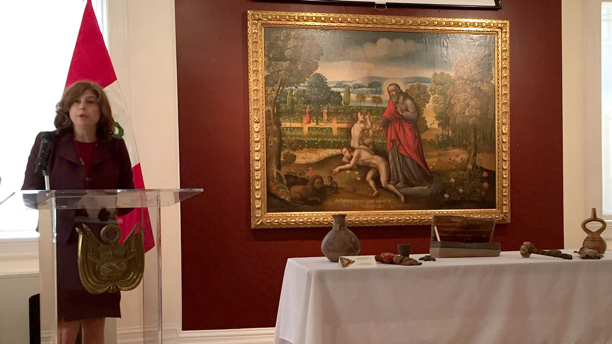 Woman stands at podium with large painting on a wall beside her and artifacts on a table